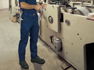 Homem com calças de trabalho azuis e sapatos de segurança verdes junto a máquina industrial bege