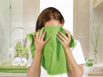 Mulher a cobrir o rosto com uma toalha verde numa casa de banho com decoração de toalhas e plantas