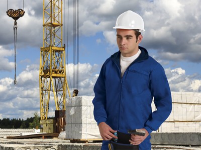 Homem com fato azul e capacete branco numa obra com grua e materiais de construção