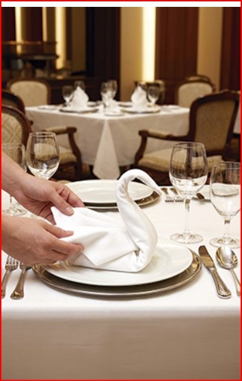 Mesa posta com toalha branca, guardanapo dobrado estilo cisne, pratos brancos com borda dourada, talheres e copos de vidro em restaurante