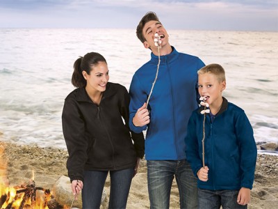 Três pessoas de casacos fleece seguram pauzinhos com marshmallows junto a fogueira na praia.