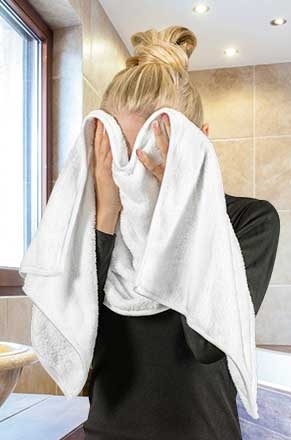 Pessoa a secar o rosto com toalha branca dentro de casa de banho com azulejos castanhos.