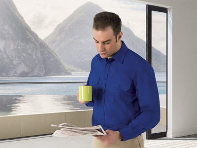 Homem com camisa azul e caneca amarela lendo jornal numa sala com vista para montanhas e lago.