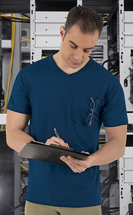 Homem com t-shirt azul com óculos no bolso segurando prancheta em ambiente com servidores