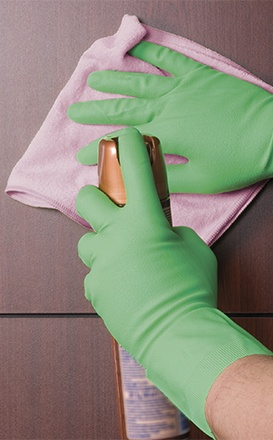 Mãos com luvas verdes segurando spray dourado e pano rosa sobre madeira