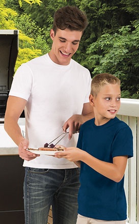 Dois rapazes com t-shirts branca e azul junto a churrasqueira numa varanda com árvores ao fundo.