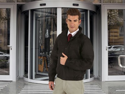 Homem com casaco preto, calças bege e gravata vermelha em frente a porta giratória de vidro