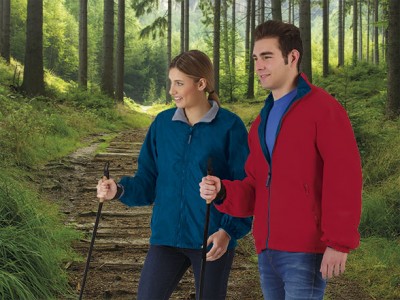 Duas pessoas vestem blusões de tecido polar azul e vermelho numa floresta, segurando bastões de caminhada