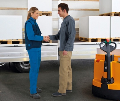 Dois trabalhadores cumprimentando-se numa área de armazenamento industrial com empilhadora elétrica amarela e caixas em paletes