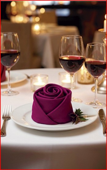 Mesa de restaurante com guardanapo roxo em forma de rosa e copos de vinho