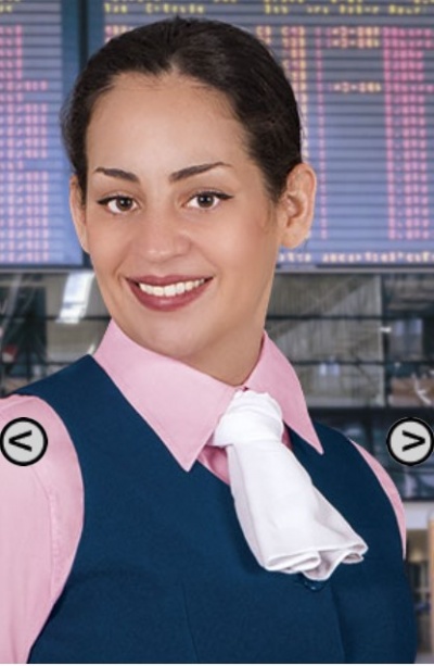 Mulher vestida com camisa rosa, colete azul escuro e lenço branco ao pescoço em ambiente com ecrãs eletrónicos.