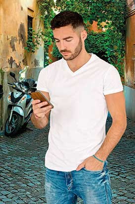 Homem com t-shirt branca e jeans segurando smartphone numa rua com motas e árvores