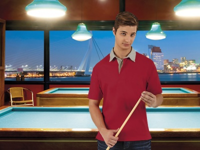 Homem jovem com polo vermelha e gola cinzenta segurando taco de bilhar em sala com mesa de bilhar e vista noturna da cidade