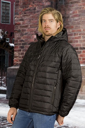 Homem com casaco acolchoado preto de capuz, fecho de correr e punhos elásticos, em cenário urbano com neve e parede de tijolos.