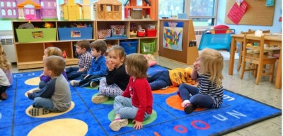 Crianças sentadas num tapete colorido numa sala de aula com mobiliário e brinquedos