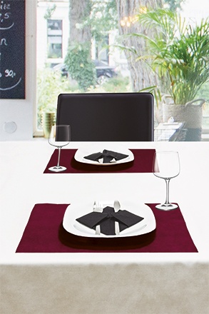 Mesa posta com pratos, guardanapos pretos, jogos americanos bordô, taças e cadeira preta junto a uma janela com plantas