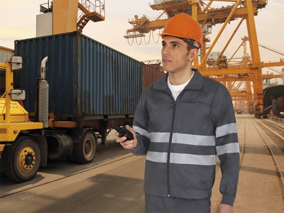 Homem com uniforme cinzento e capacete laranja num cais industrial com camião e contentor