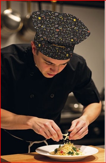 Cozinheiro de chapéu preto com padrões coloridos a preparar prato de massa numa cozinha profissional
