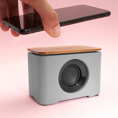 Coluna de som portátil cinza com topo de madeira e telemóvel preto sobre fundo rosa