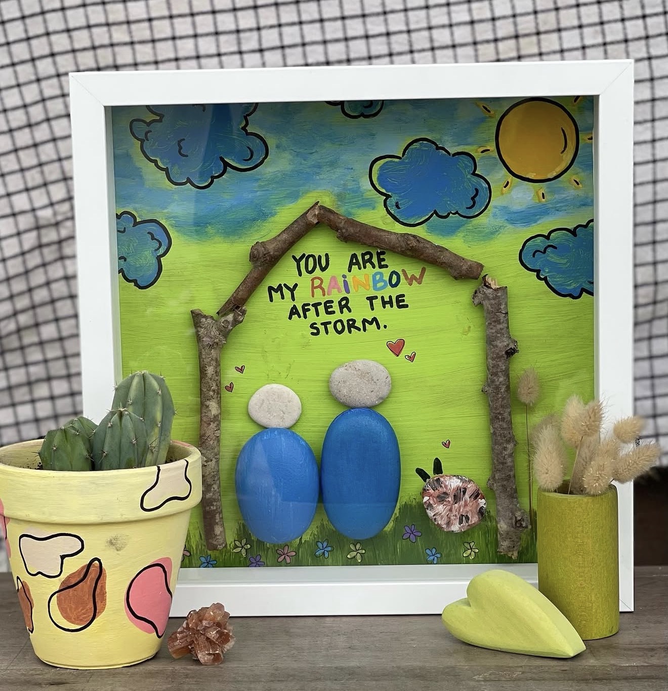 Quadro decorativo colorido com pedras, ramos secos e texto inspirador, ao lado de vasos com cactos e ramos secos.