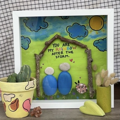 Quadro decorativo colorido com pedras, ramos secos e texto inspirador, ao lado de vasos com cactos e ramos secos.