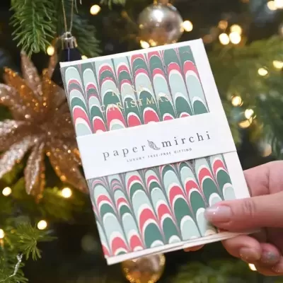 Pacote de cartão festivo com padrão colorido e texto dourado, segurado em frente a uma árvore de Natal com luzes.