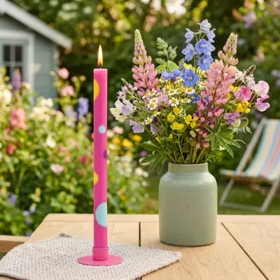 Vela rosa com pontos coloridos ao lado de vaso com flores num jardim