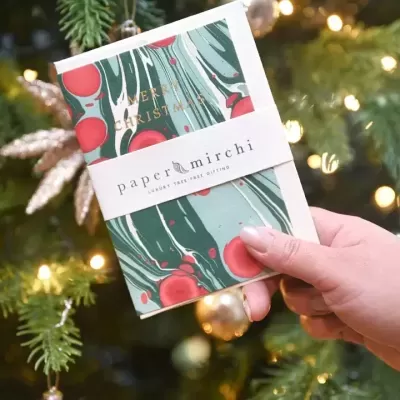 Cartão de Natal colorido com a frase MERRY CHRISTMAS e marca papermirchi, em frente a uma árvore de Natal