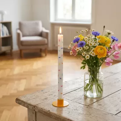 Vela branca com pontos coloridos, em suporte amarelo, numa mesa de madeira rústica ao lado de jarro com flores coloridas num ambiente interior iluminado