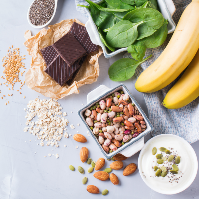 Alimentos saudáveis diversos como chocolate, espinafre, banana, feijões, amêndoas, sementes, aveia e iogurte em recipientes e espalhados sobre uma superfície clara.