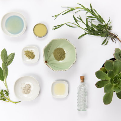 Tigelas com pós coloridos, líquidos, plantas verdes e garrafa de vidro sobre fundo branco.