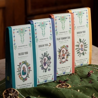 Quatro caixas de chá floral com design oval e ervas sobre tecidos verdes com secas de frutas.