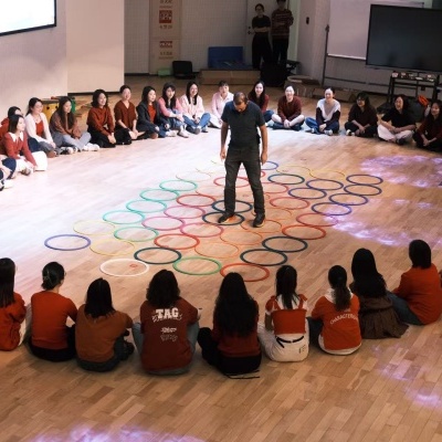 Adultos sentados em círculo com argolas coloridas no chão e pessoa em pé no centro.