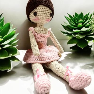 Boneca de croché rosa e castanha sentada entre plantas verdes