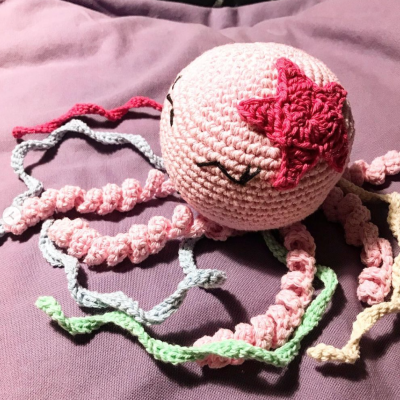 Boneco de crochê rosa em forma de polvo com tentáculos coloridos sobre pano roxo
