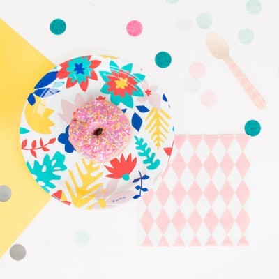 Donut rosa com granulados num prato de papel floral sobre superfície branca com confetes e papel decorativo.