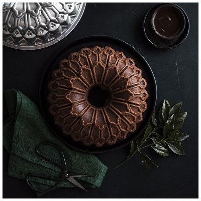 Bolo com padrão geométrico em prato preto, pano verde, tesoura e folhas sobre mesa escura