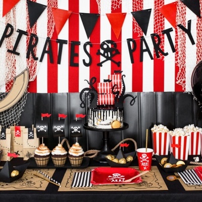 Mesa de festa decorada com tema de pirata, incluindo bolo, cupcakes, pipoca, balões e toalha preta.