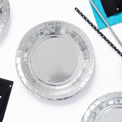 Pratos de papel prateados, guardanapos azuis e pretos com pontos brancos, palhinhas e copos metálicos em mesa branca