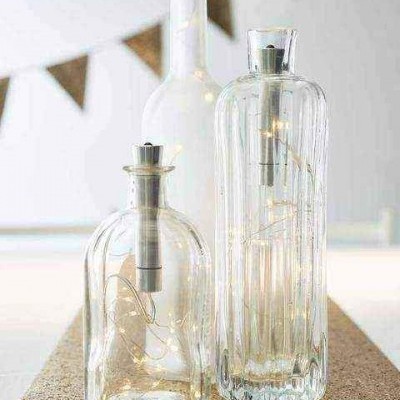 Duas garrafas de vidro com luzes LED em superfície de cortiça e bandeirolas ao fundo