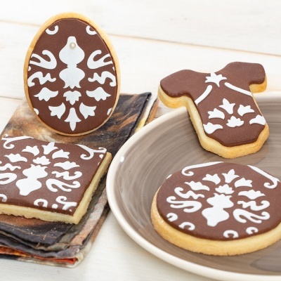 Bolachas decoradas em chocolate com padrões florais brancos, formas oval, quadrada e roupa, sobre travessa e panos