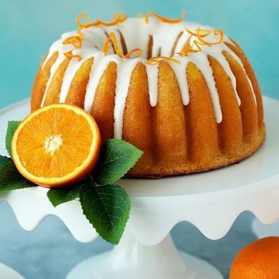 Bolo bundt com cobertura branca e casca de laranja decorativa sobre suporte branco com metade de laranja e folhas verdes