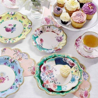 Pratos e chávenas de porcelana decorados com flores coloridas e bordas onduladas em mesa branca com cupcakes e flores
