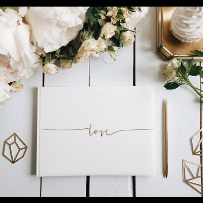 Caderno branco com palavra 'love', caneta dourada, flores brancas e adornos geométricos dorados numa mesa de madeira branca