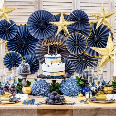 Mesa decorada para festa com bolo, flores, uvas, estrelas douradas e leques azuis ao fundo