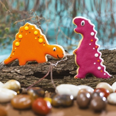 Biscoitos decorados em forma de dinossauros coloridos sobre madeira com pedras ao redor