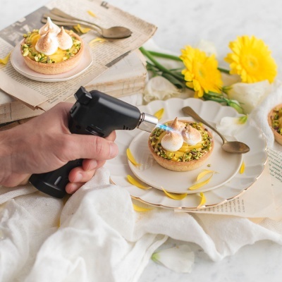 Mão a usar maçarico para dourar suspiros em tarte com pistácios em prato branco com flores amarelas