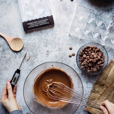 Utensílios e ingredientes para fazer chocolate com tigela de vidro, batedor metálico, colher de madeira, molde de silicone e gotas de chocolate