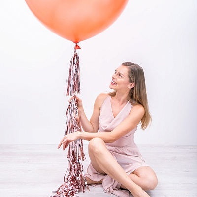 Mulher com vestido rosa claro segura balão laranja com fitas prateadas sentada no chão