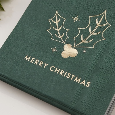 Guardanapo de papel verde com mensagem 'MERRY CHRISTMAS' e desenho dourado de folhas de azevinho.